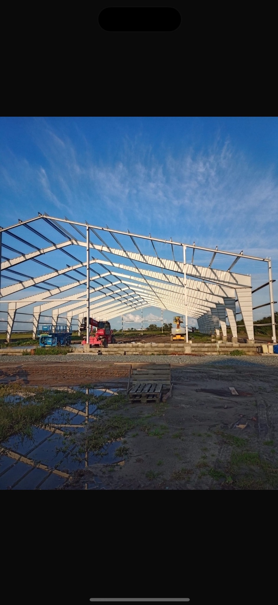 Stalowa konstrukcja hali w trakcie budowy, widoczne podnośniki koszowe i palety, na pierwszym planie kałuża odbijająca niebo.