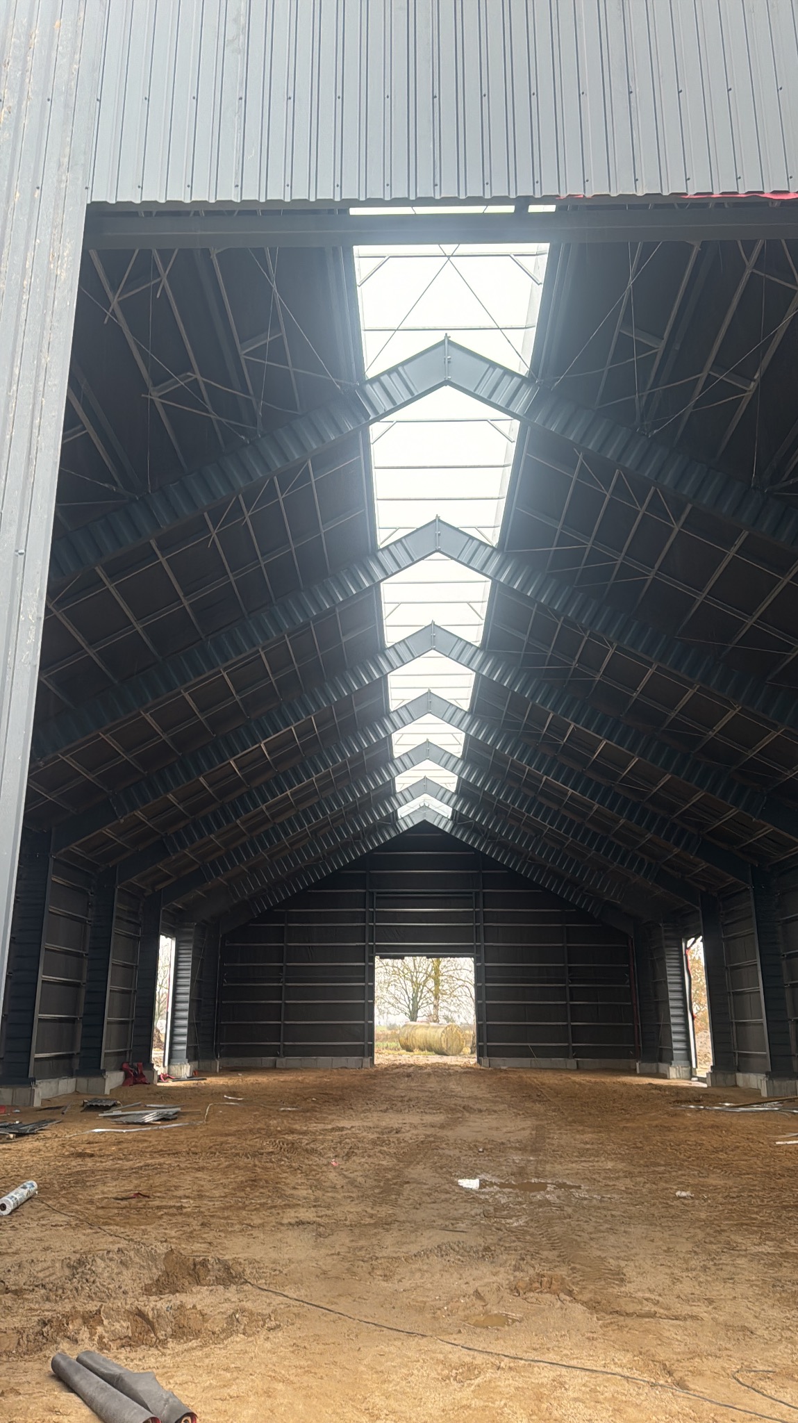 Wnętrze nowo wznoszonej hali stalowej z widoczną konstrukcją dachu i świetlikami dachowymi, surowy grunt przygotowany pod wylewkę.