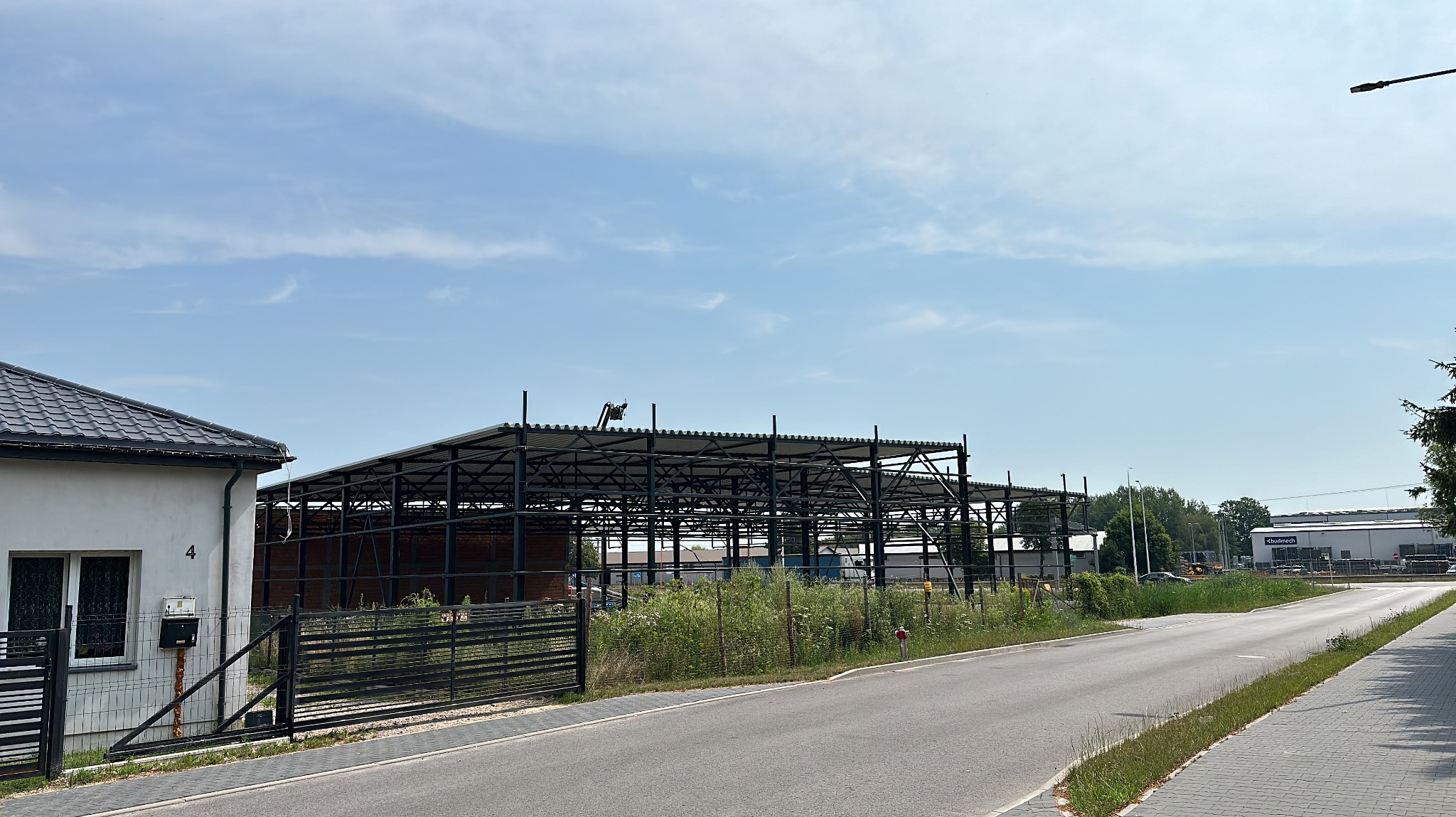 Wznoszona konstrukcja stalowa hali na tle błękitnego nieba, widoczna zza ogrodzenia posesji z domem jednorodzinnym.