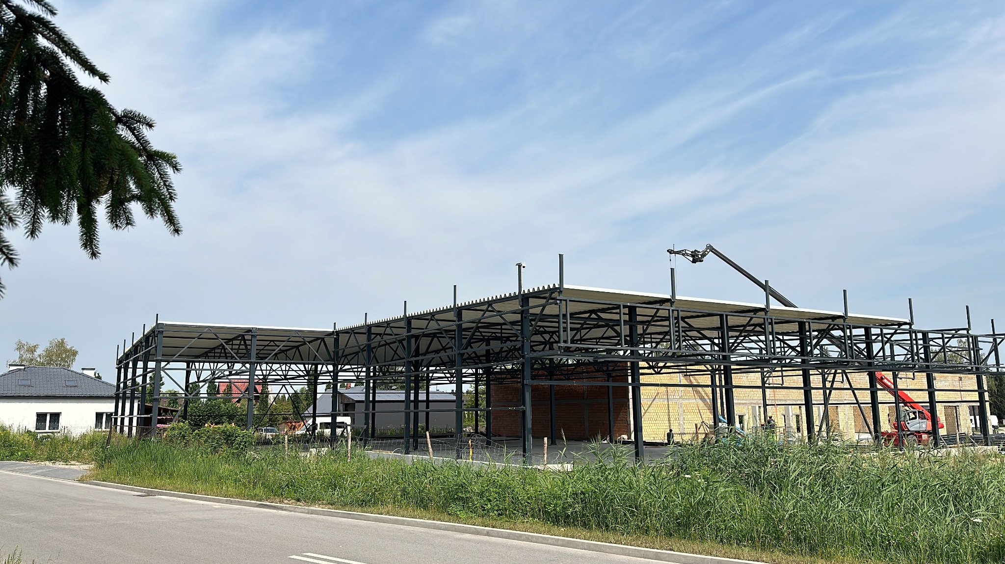 Konstrukcja stalowa hali w trakcie budowy, widoczny żuraw teleskopowy, częściowo wymurowane ściany z żółtej cegły, w tle domy jednorodzinne i błękitne niebo z chmurami.