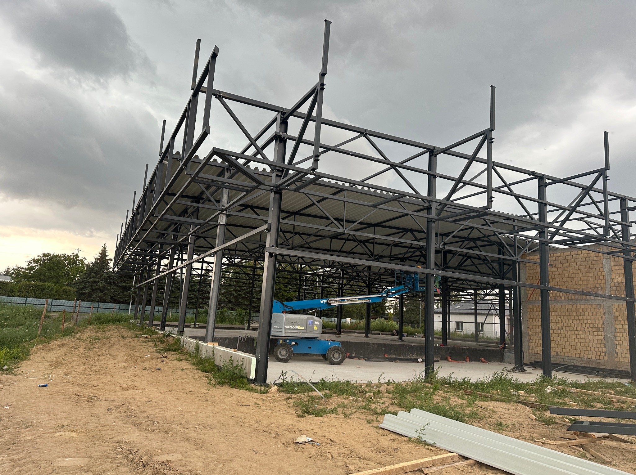 Konstrukcja stalowa hali w trakcie budowy, widoczny podnośnik koszowy i częściowo wzniesione ściany z cegły silikatowej, pochmurne niebo w tle.