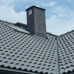 Szary komin z kratką wentylacyjną wystający ponad dachówkę w odcieniach szarości i błękitne niebo z chmurami w tle.