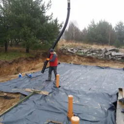 Pracownik w czerwonej kamizelce układa czarną folię izolacyjną na fundamencie domu, widoczne rury wentylacyjne i fragment wykopu.