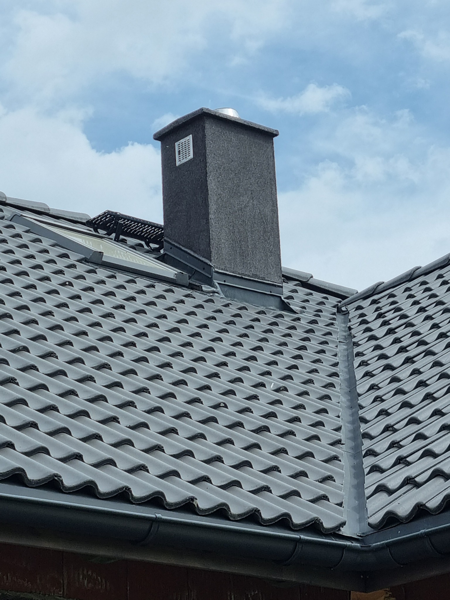 Szary komin z kratką wentylacyjną wystający ponad dachówkę w odcieniach szarości i błękitne niebo z chmurami w tle.