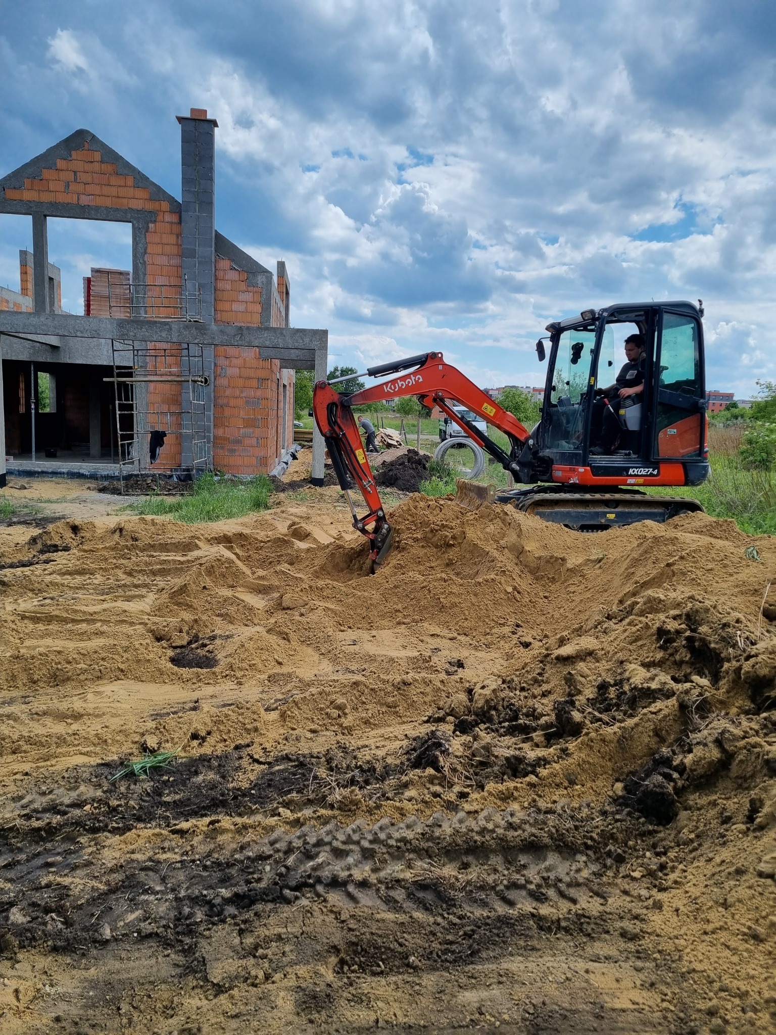 Mała koparka Kubota na placu budowy, w tle surowy budynek z czerwonej cegły w trakcie wznoszenia ścian, widoczne rusztowania.