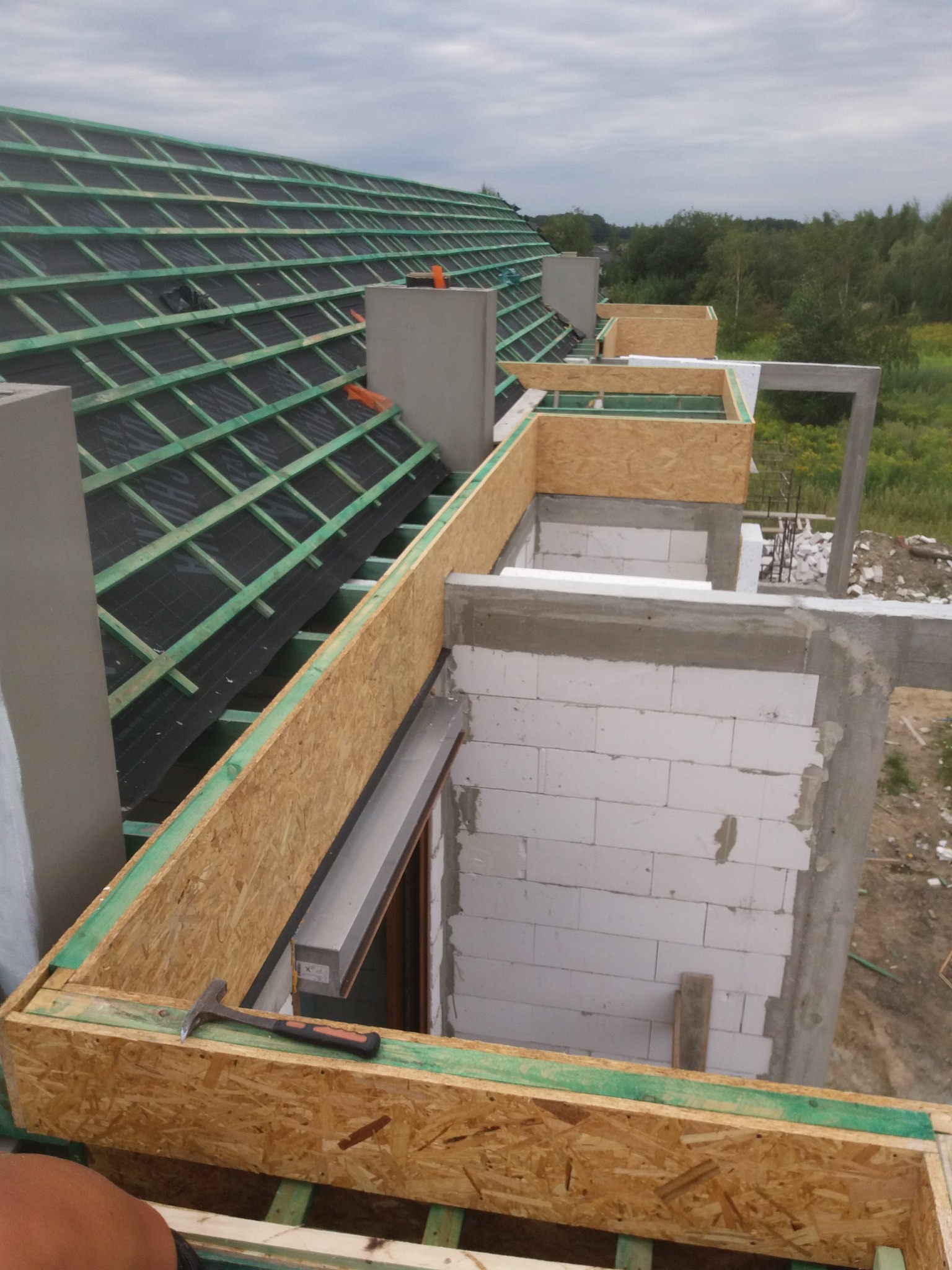 Wznoszenie ścian z białych bloczków na nowym budynku, widoczne elementy konstrukcji dachu z łatami i membraną, oraz fragment drewnianej szalówki, na pierwszym planie leży młotek.