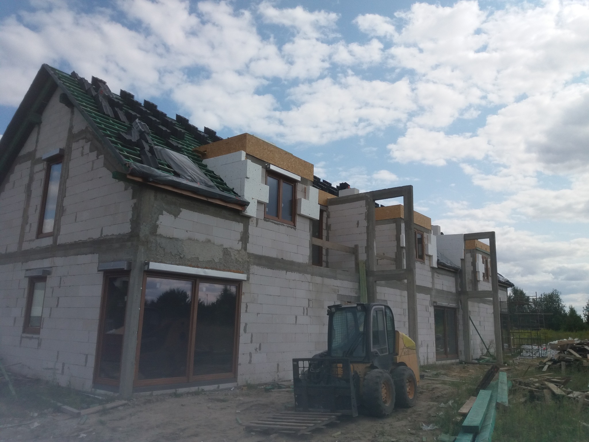 Niedokończony dom murowany z białych pustaków, widoczny częściowo położony dach, okna z brązowymi ramami i żółta ładowarka teleskopowa na placu budowy.