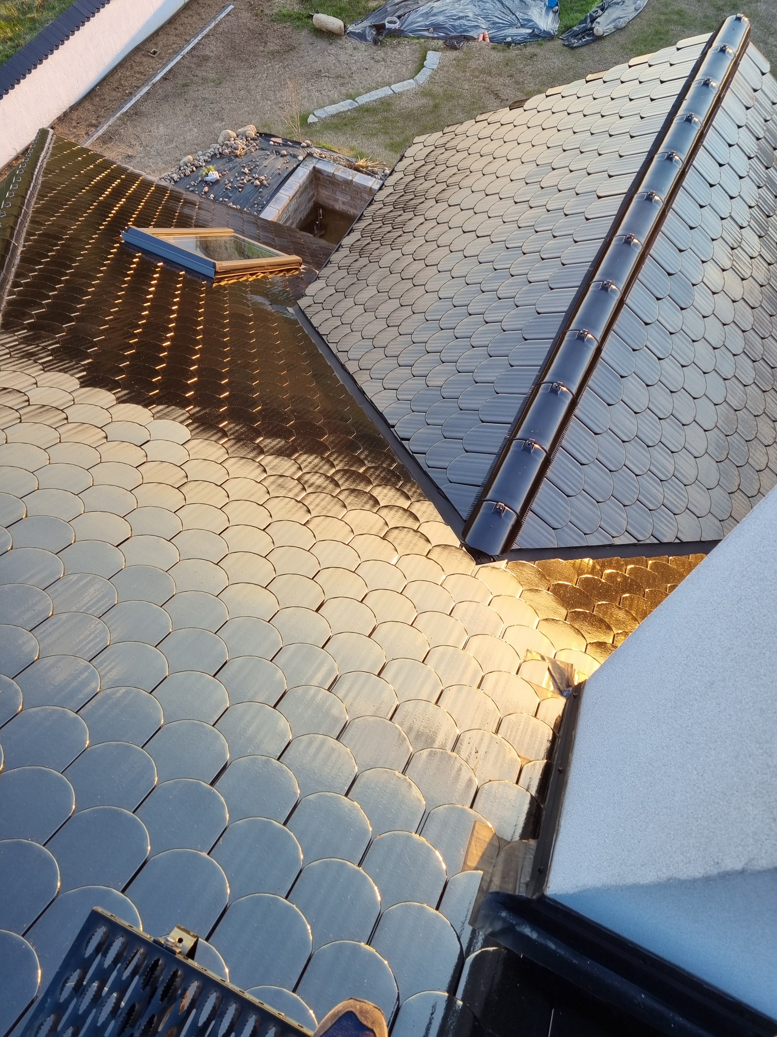 Widok z góry na dach pokryty błyszczącymi, półokrągłymi dachówkami w dwóch odcieniach szarości, z zamontowanym oknem dachowym i systemem odprowadzania wody. Część elewacji budynku widoczna w prawym...