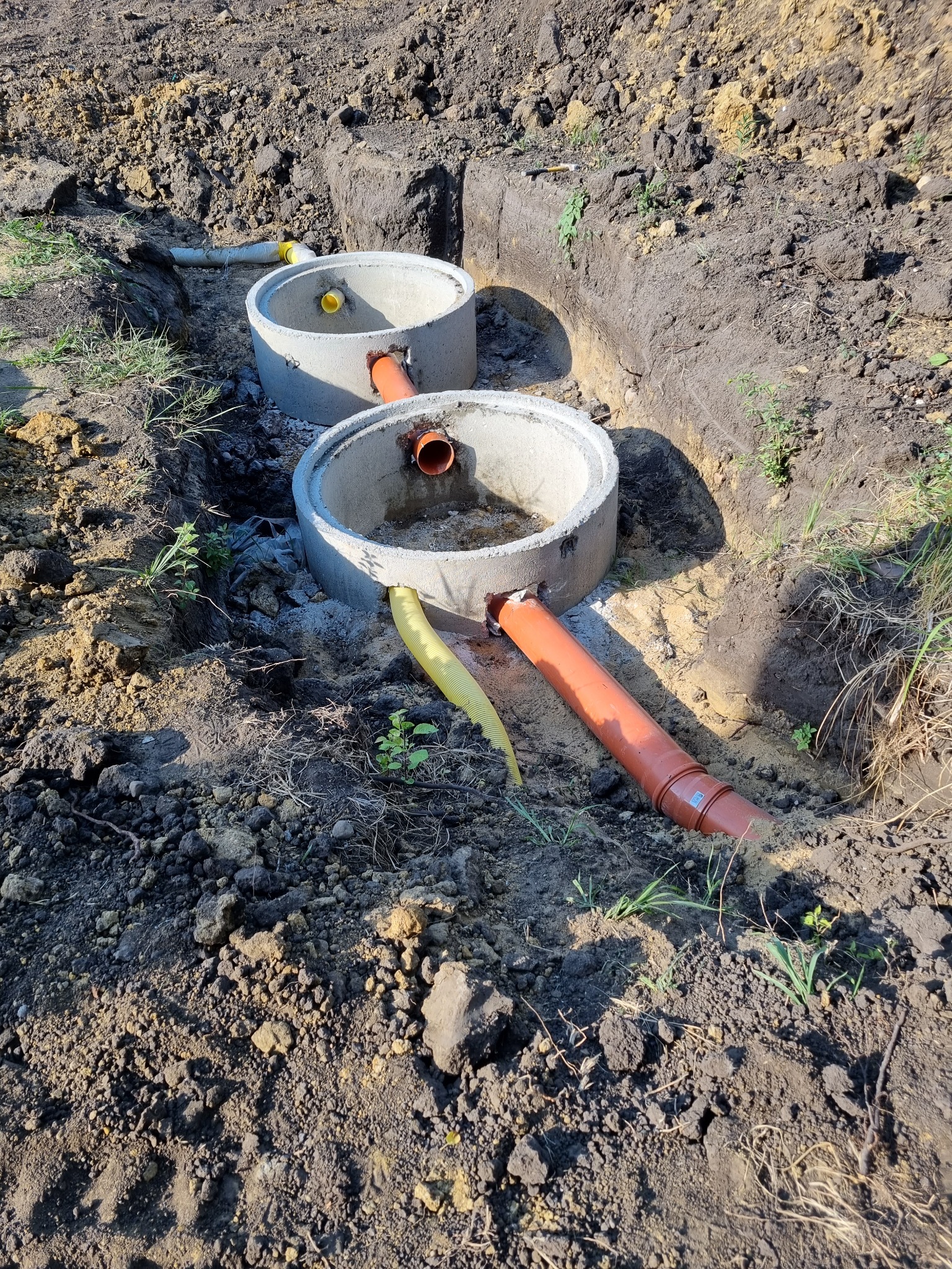 Dwa betonowe kręgi studni połączone rurami z pomarańczowego PCV i żółtą karbowaną rurą, umieszczone w wykopanym rowie w ziemi.