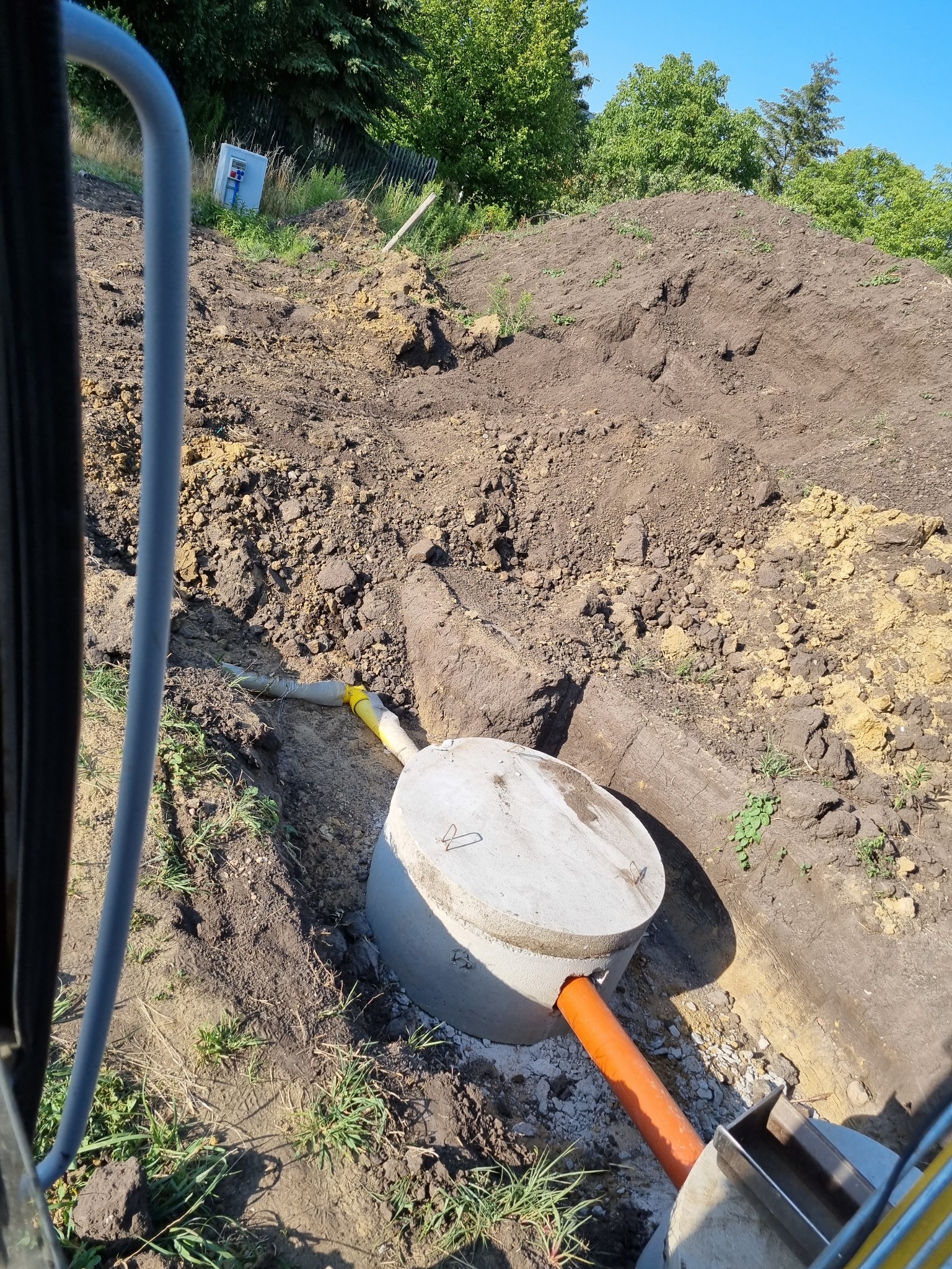 Montaż betonowego zbiornika podziemnego z podłączonymi rurami kanalizacyjnymi w wykopie, widok z góry z perspektywy maszyny budowlanej.