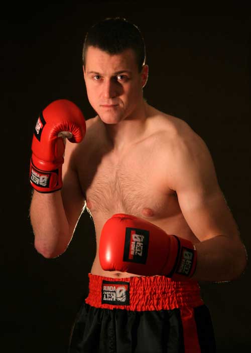 Młody mężczyzna w czerwonych rękawicach bokserskich i spodenkach do kickboxingu, pozujący na czarnym tle w studiu.