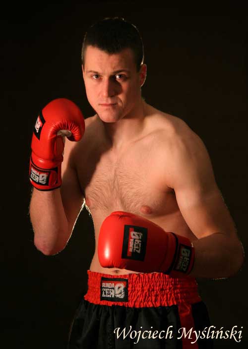 Młody mężczyzna w czerwonych rękawicach bokserskich i czarno-czerwonych spodenkach, pozujący na czarnym tle.