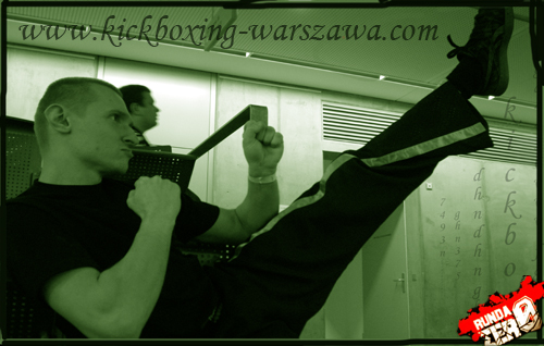 Mężczyzna w czarnym stroju siedzi na krześle, wykonując kopnięcie nogą do góry, w tle widoczna ściana z napisanymi literami i adres strony internetowej