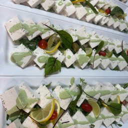 TASTY CATERING Tomasz Szmidt - Elegancko ułożone na białych talerzach kwadratowe przekąski z białego sera polane zielonym sosem, udekorowane zieleniną, pomidorkami i plasterkami cytryny.