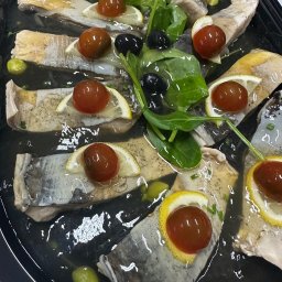 TASTY CATERING Tomasz Szmidt - Śledzie w oleju, udekorowane cytryną, pomidorkami cherry, oliwkami i świeżym szpinakiem, ułożone na czarnym talerzu. Gotowa potrawa cateringowa.