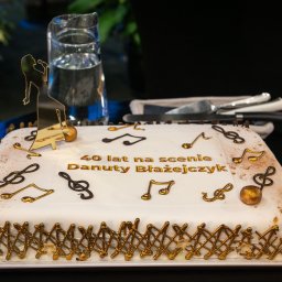 TASTY CATERING Tomasz Szmidt - Tort urodzinowy z napisem '40 lat na scenie Danuty Błażejczyk', nutami i figurką piosenkarki, na białym talerzu, obok dzbanek z wodą i sztućce.