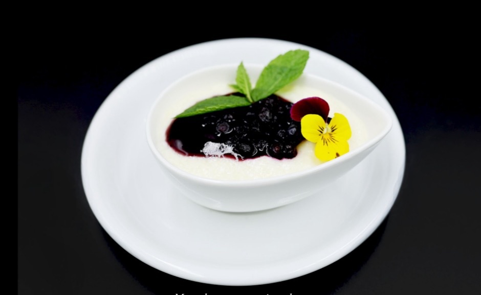 Elegancko podany deser: delikatny krem z sosem jagodowym, udekorowany listkiem mięty i jadalnym kwiatem bratka, na białym talerzu i czarnym tle.