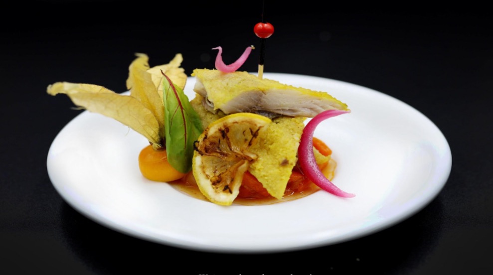 Wykwintna kompozycja na białym talerzu: filet z ryby z grilla, physalis, karmelizowana cytryna i dekoracyjne pikle na czarnym tle.