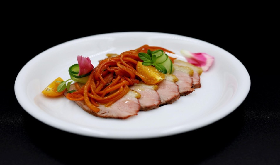 Elegancko podane plastry pieczonego mięsa z karmelizowaną marchewką julienne, pomarańczą, ogórkiem i płatkami róż na białym talerzu, czarne tło.