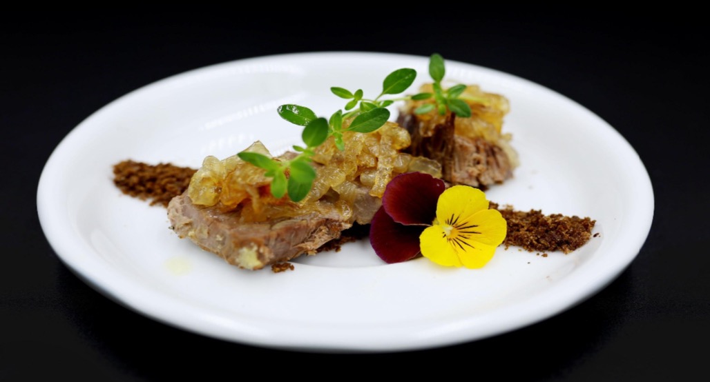 Elegancko podane danie mięsne na białym talerzu z sosem cebulowym, świeżym oregano i kwiatami bratka na czarnym tle.