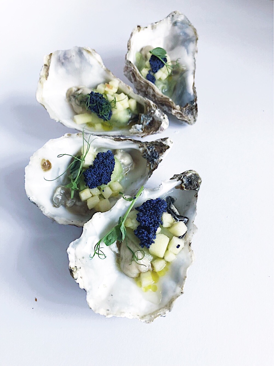 Trzy otwarte ostrygi na białym tle, wypełnione tartym jabłkiem, kawiorem i dekorowane pędami groszku.