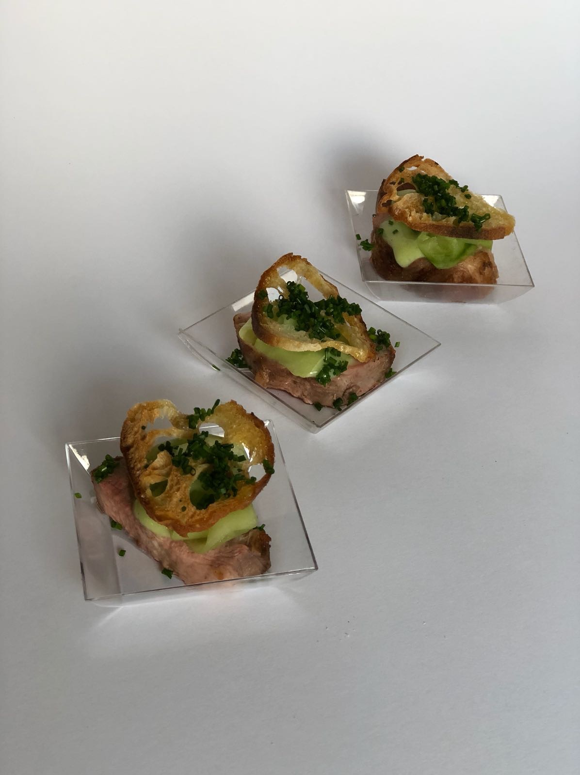 Trzy miniaturowe kanapki z rostbefem, ogórkiem i grzanką z dziurami, posypane szczypiorkiem, ułożone na przezroczystych kwadratowych tackach na białym tle.