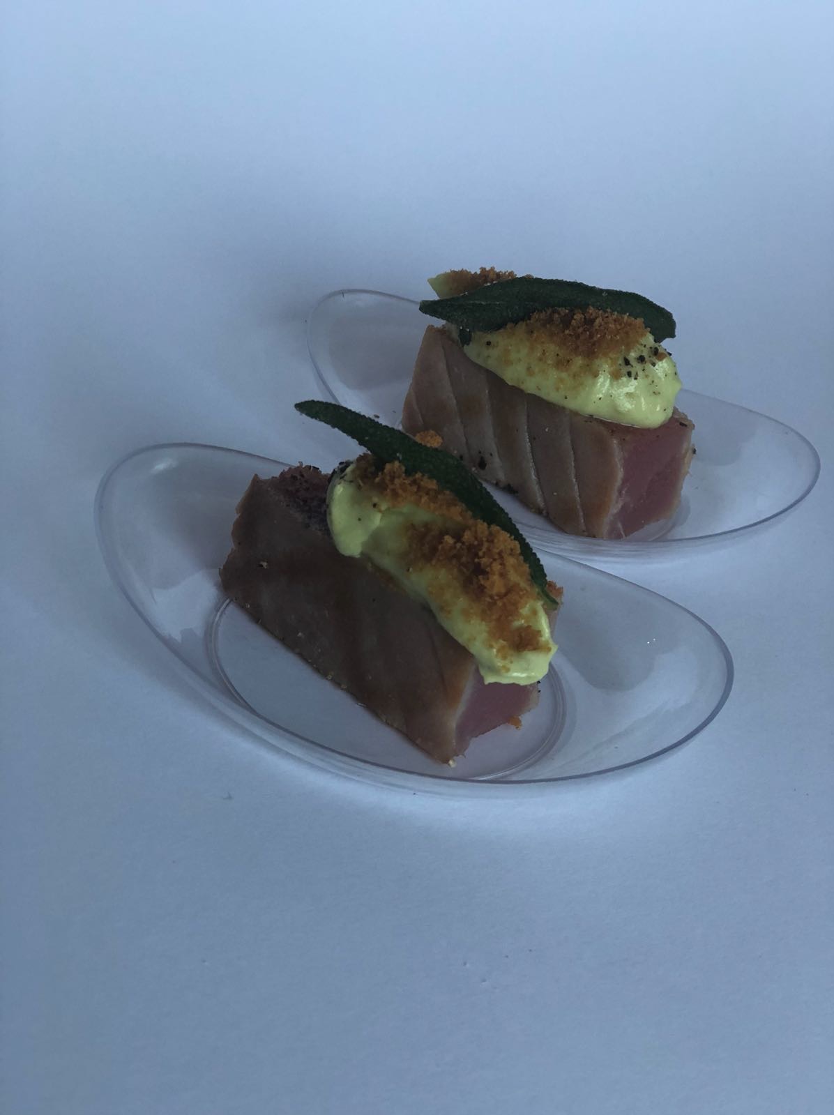 Dwa kawałki tuńczyka na przezroczystych łyżkach, polane kremowym sosem, posypane brązowymi okruchami i udekorowane listkiem szałwii.