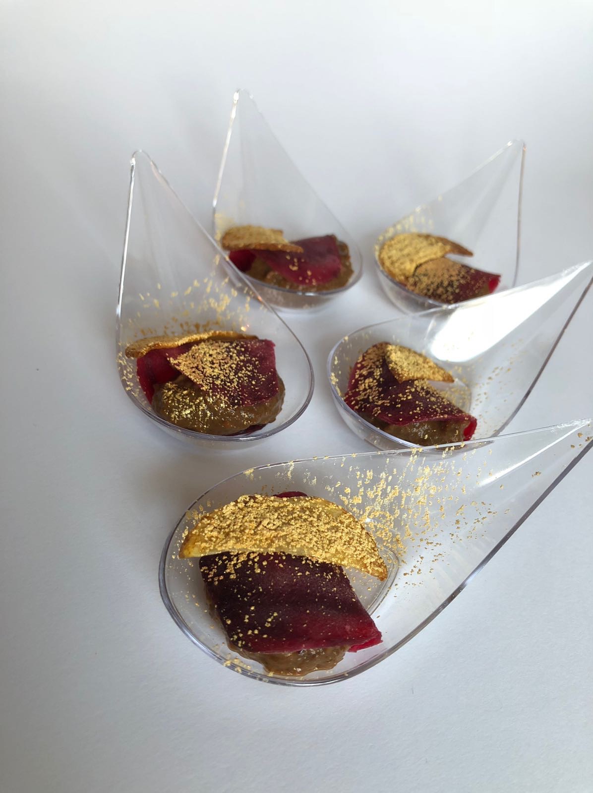 Eleganckie finger food: pięć przezroczystych łyżeczek z ciemną pastą, burakiem i kruchym ciastkiem posypanym złotym pyłem, ułożone na białym tle.