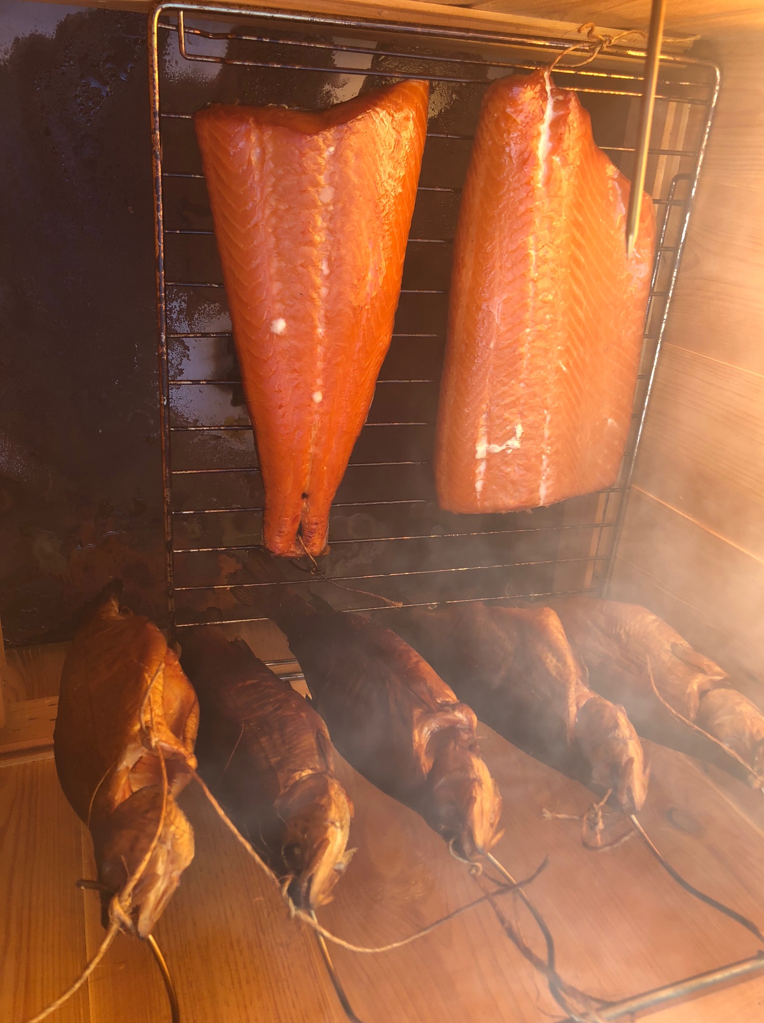 Wędzone filety z łososia i wędzone sumy w wędzarni z metalowym rusztem, widoczne delikatne smugi dymu.