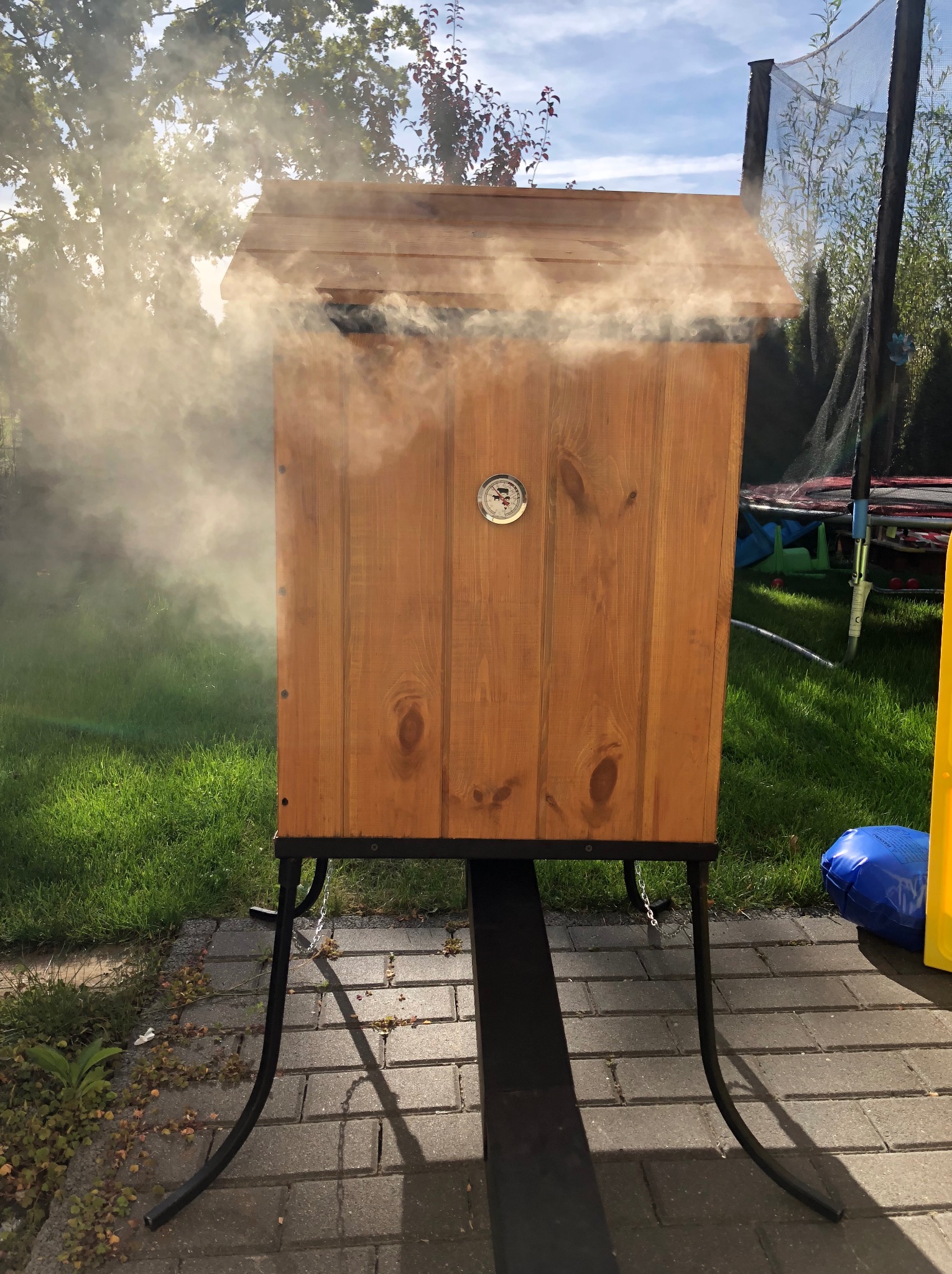 Drewniana wędzarnia ogrodowa na metalowym stelażu, z widocznym dymem wydobywającym się z górnej części, na tle trawnika i trampoliny.