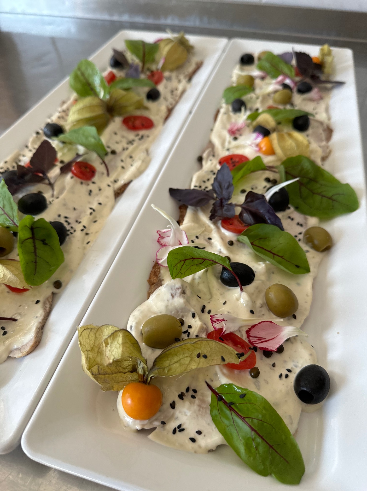 Dwa prostokątne talerze z pasztetem cielęcym, dekorowane oliwkami, pomidorkami cherry, miechunką peruwiańską i listkami ziół, posypane czarnym sezamem.