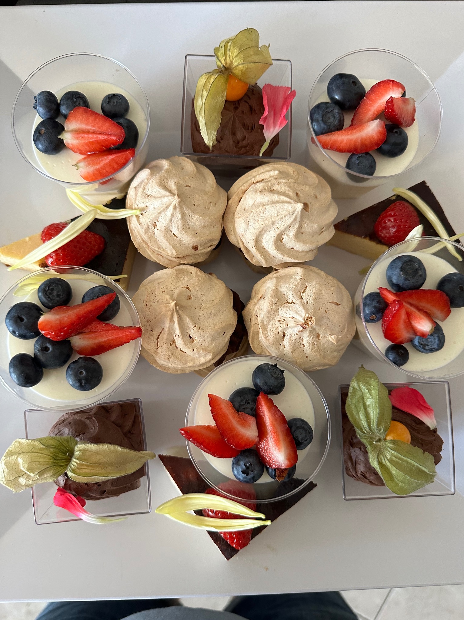 Ekskluzywny zestaw deserów: kremowe desery z borówkami i truskawkami w przezroczystych pucharkach, czekoladowe musy z miechunką peruwiańską i płatkami kwiatów oraz bezy na białym tle.