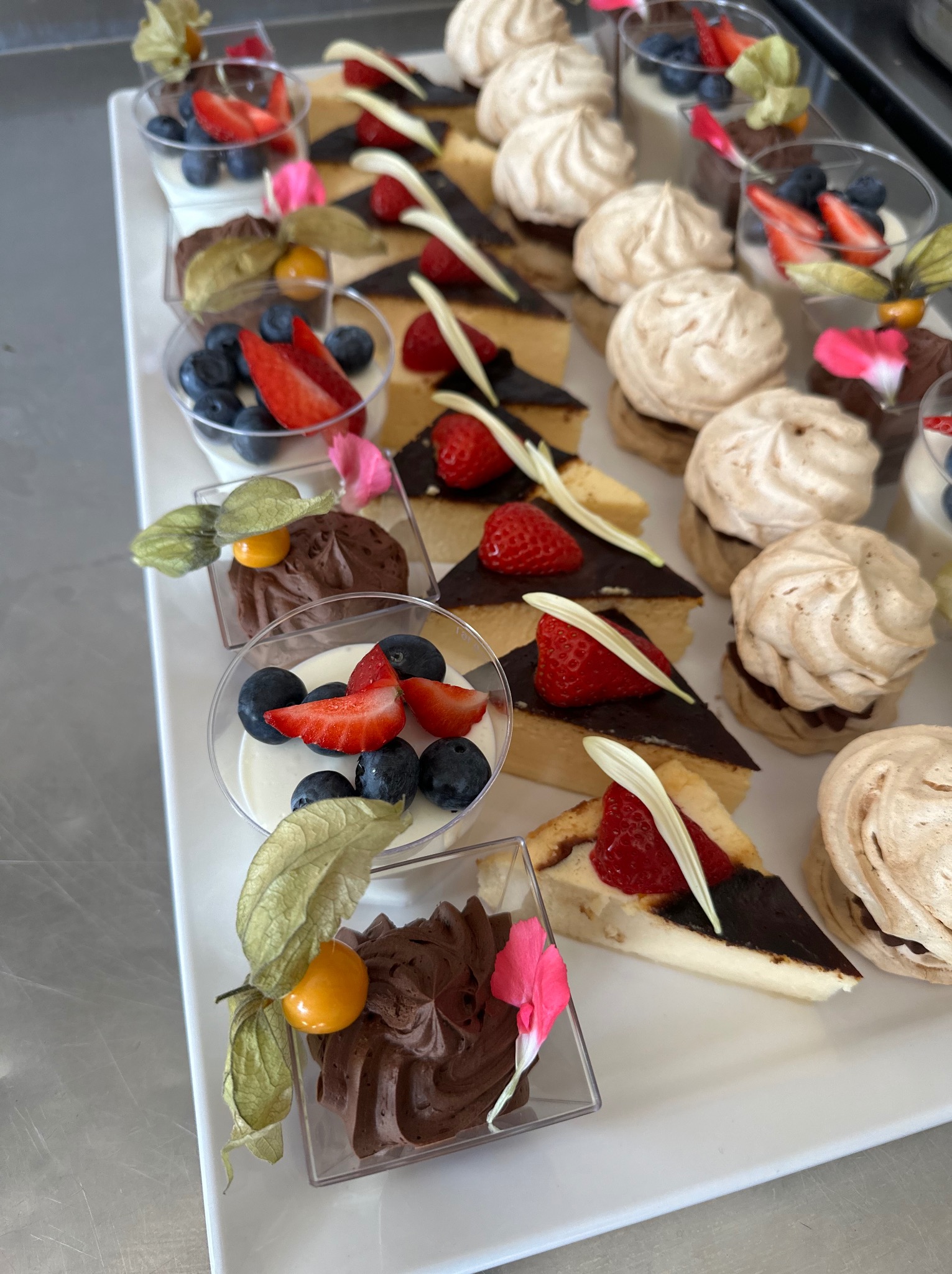 Elegancki wybór deserów: mini bezy, krem czekoladowy w kubeczkach, sernik z polewą czekoladową i deser jogurtowy z borówkami i truskawkami, udekorowane physalis i płatkami kwiatów, ułożone...