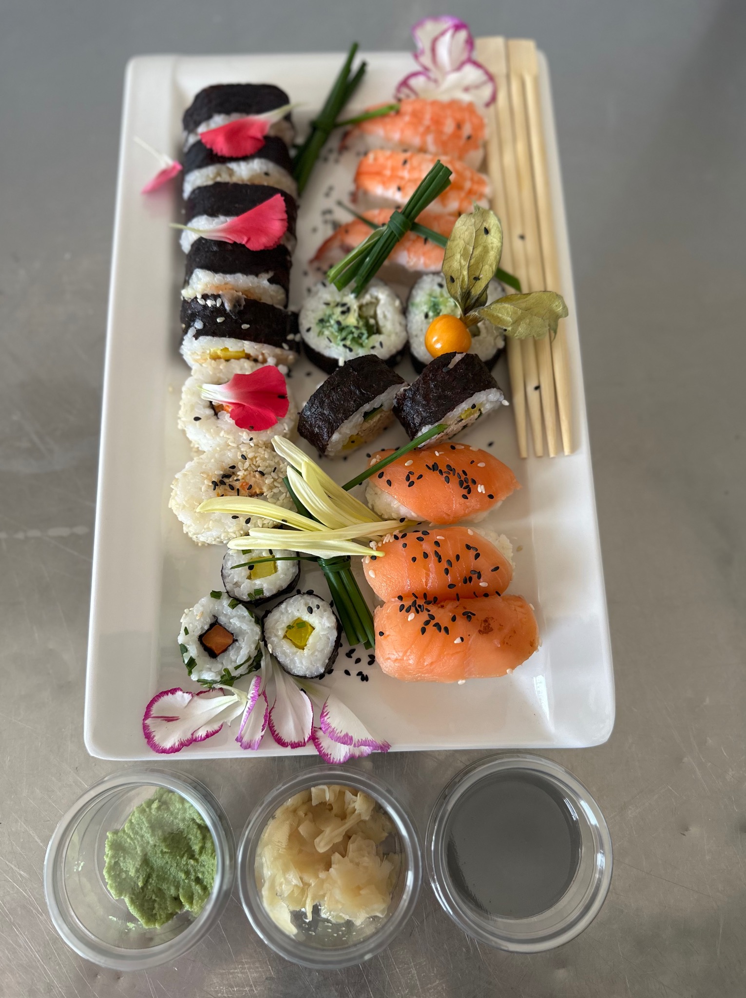 Elegancko podany zestaw sushi na białym prostokątnym talerzu, udekorowany kwiatami, z dodatkami wasabi, imbiru i sosu sojowego w przezroczystych miseczkach.