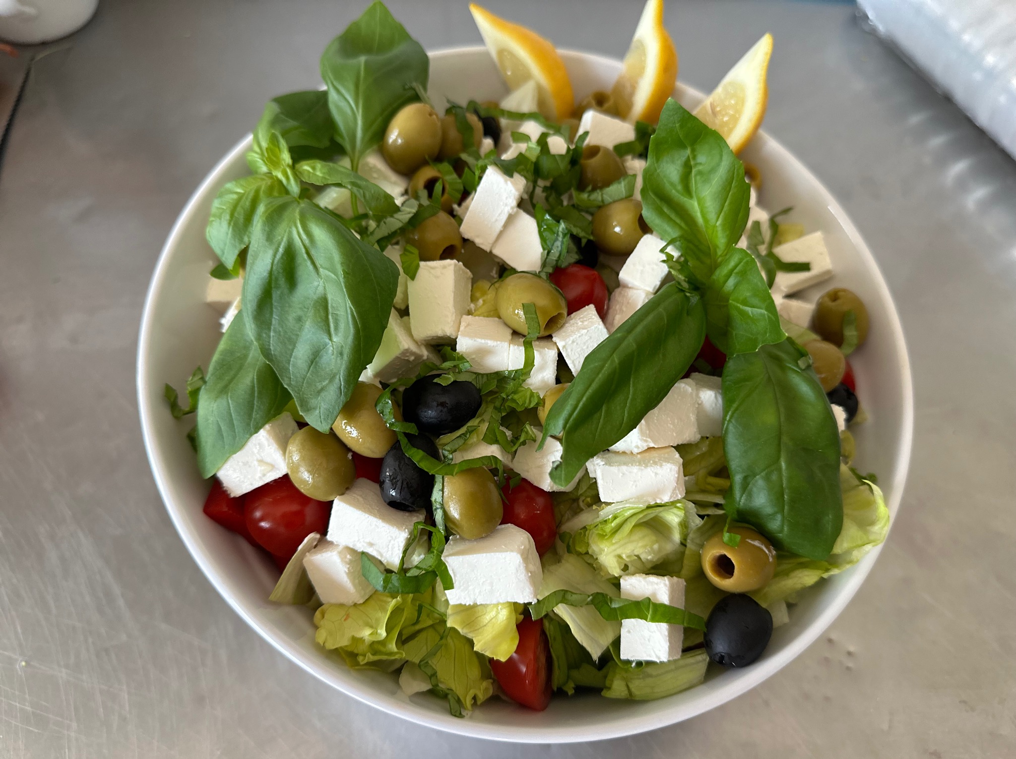 Świeża sałatka grecka w białej misce, z kostkami sera feta, oliwkami zielonymi i czarnymi, pomidorkami cherry, bazylią i plasterkami cytryny, na jasnym blacie.