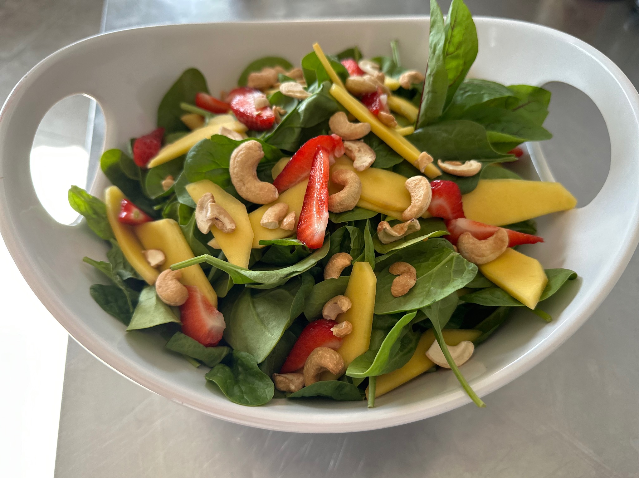 Sałatka ze szpinaku, mango, truskawek i orzechów nerkowca w białej misce z uchwytami.