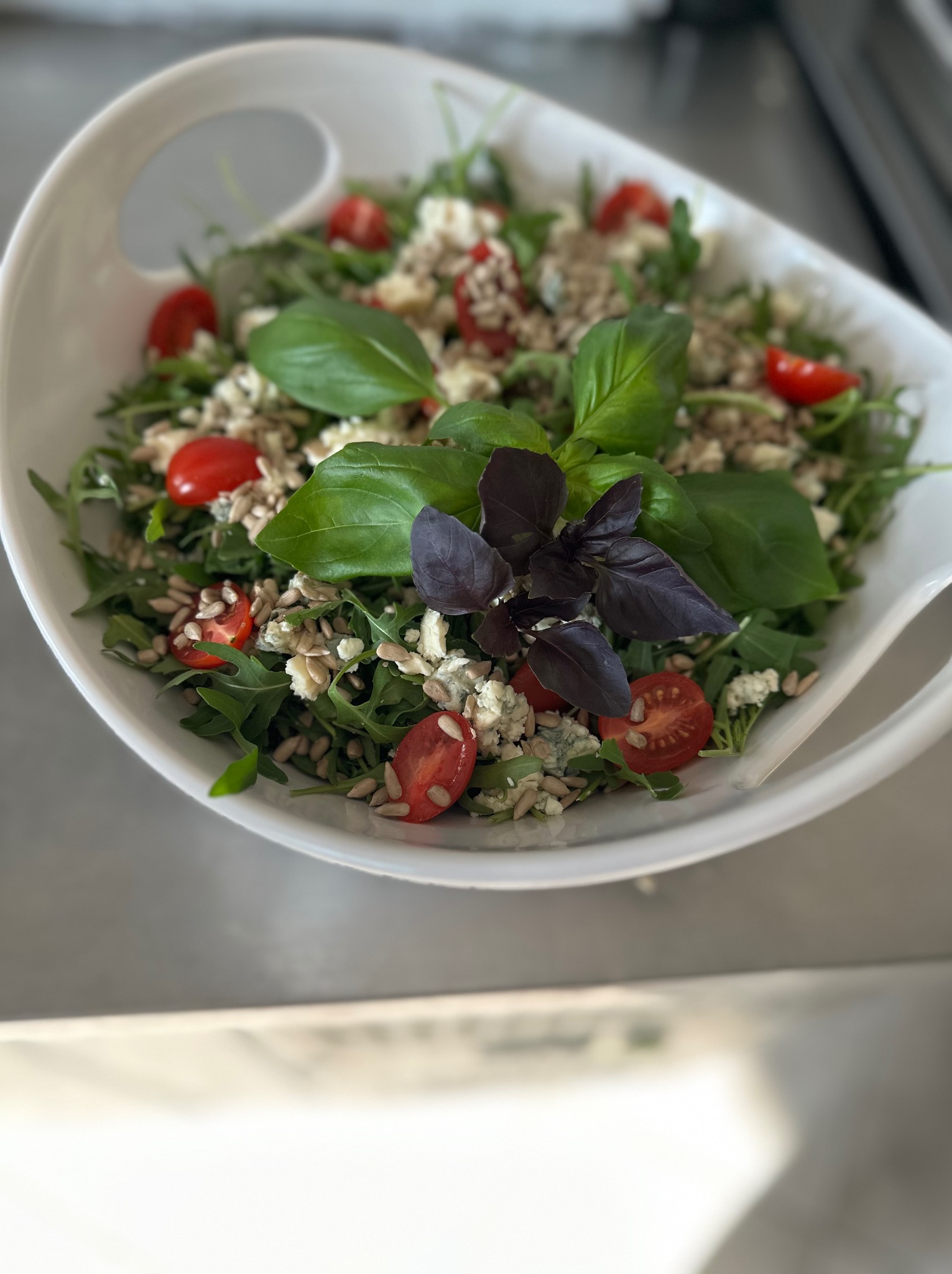 Świeża sałatka z rukoli, pomidorków cherry, sera pleśniowego i prażonych ziaren słonecznika, udekorowana świeżą bazylią w białej misce.