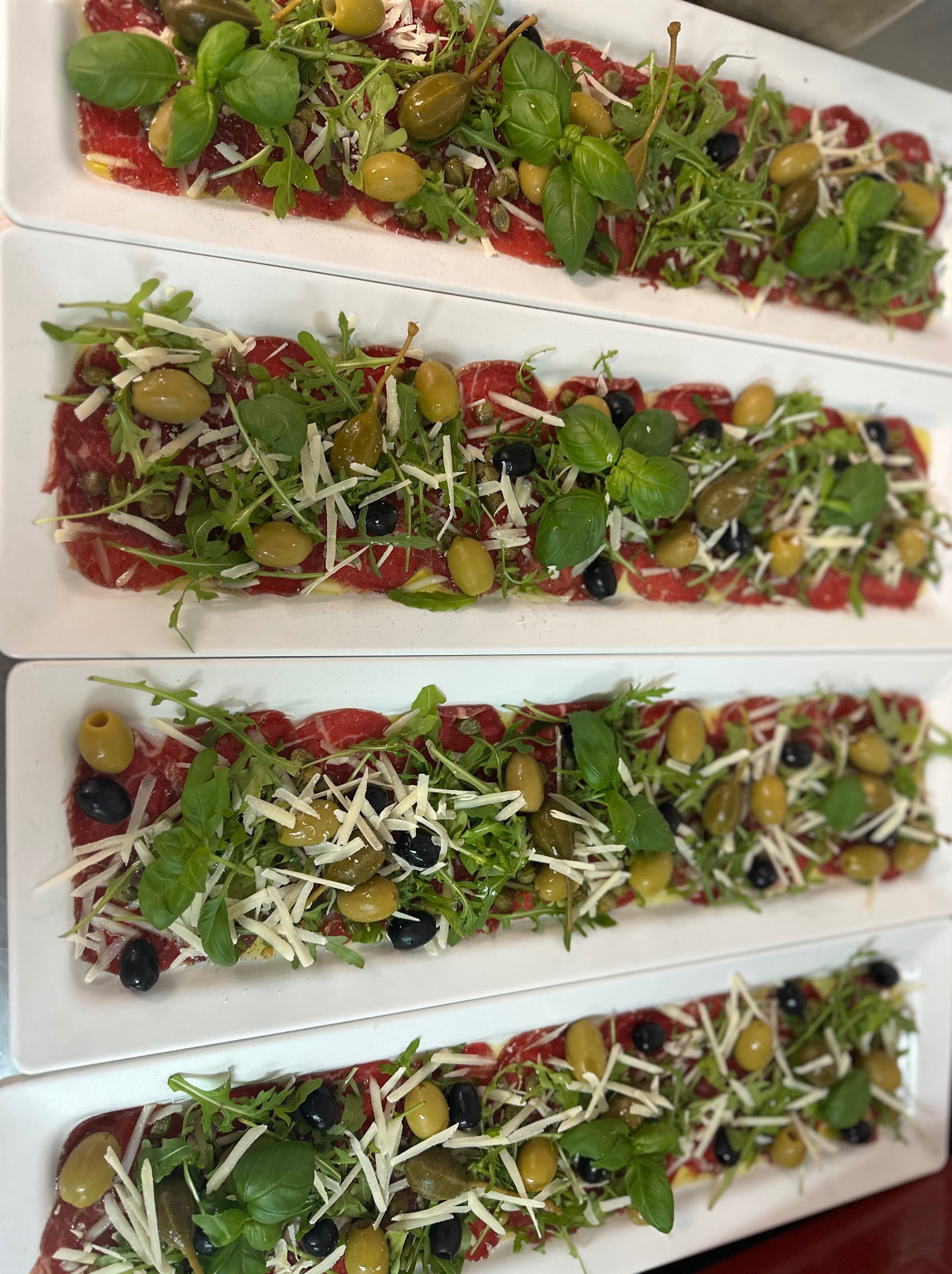Elegancko podane carpaccio wołowe na białych prostokątnych talerzach, udekorowane rukolą, kaparami, oliwkami i bazylią. Widok z góry, ostre światło.