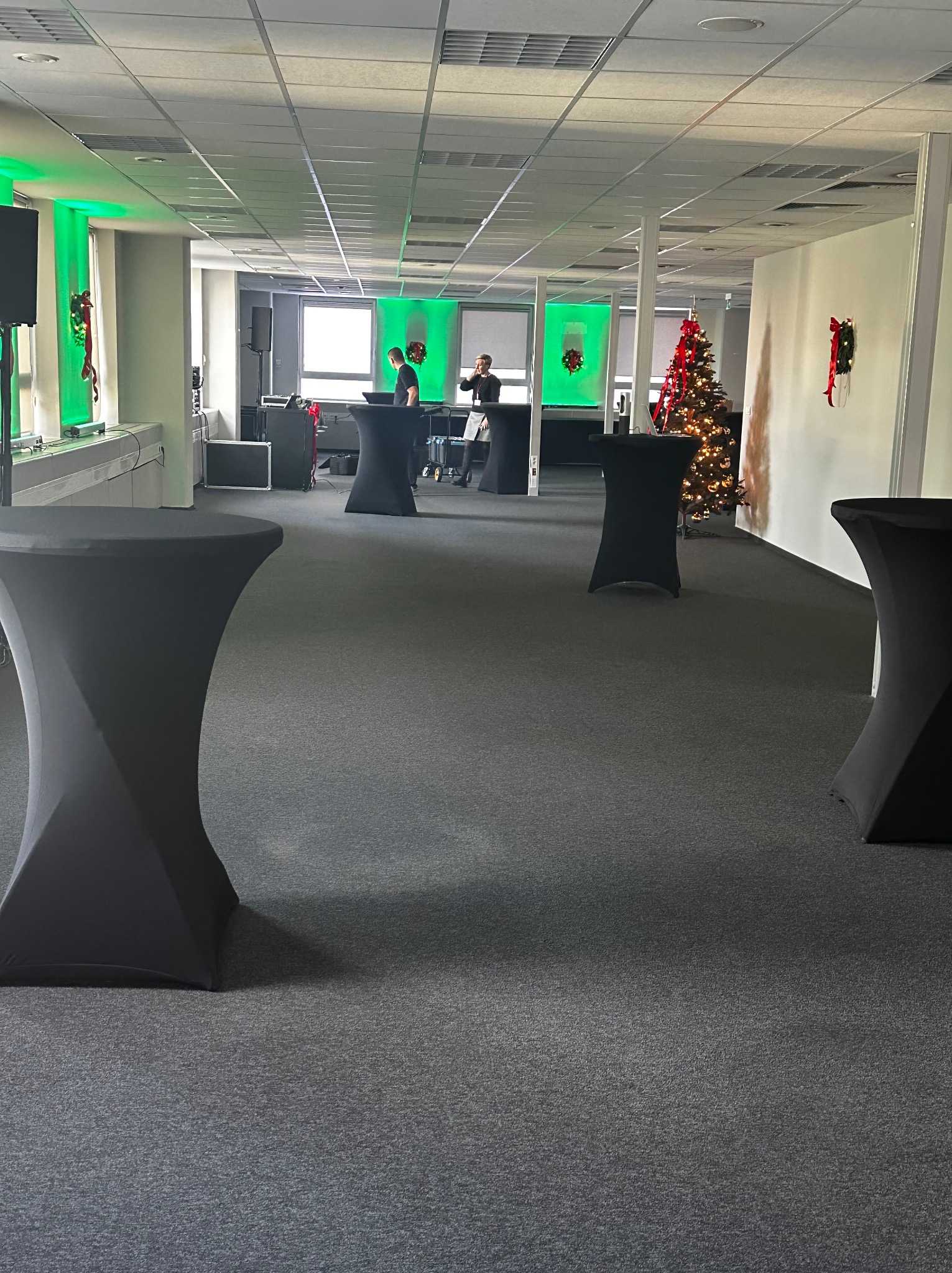 Biuro z dekoracjami świątecznymi i czarnymi stolikami koktajlowymi, przygotowane do firmowego przyjęcia. W tle osoby przy stanowisku DJ-skim i choinka.