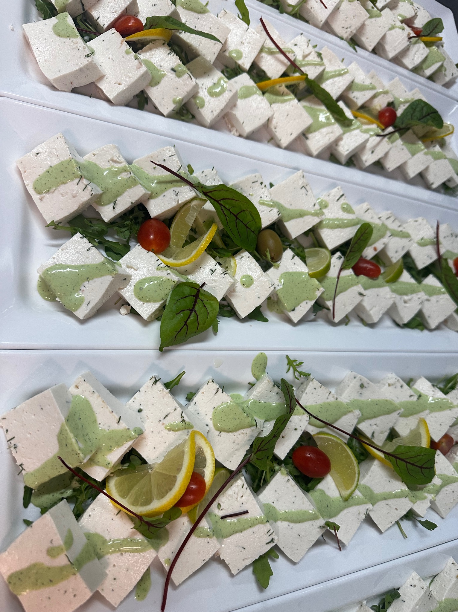 Elegancko ułożone na białych talerzach kwadratowe przekąski z białego sera polane zielonym sosem, udekorowane zieleniną, pomidorkami i plasterkami cytryny.
