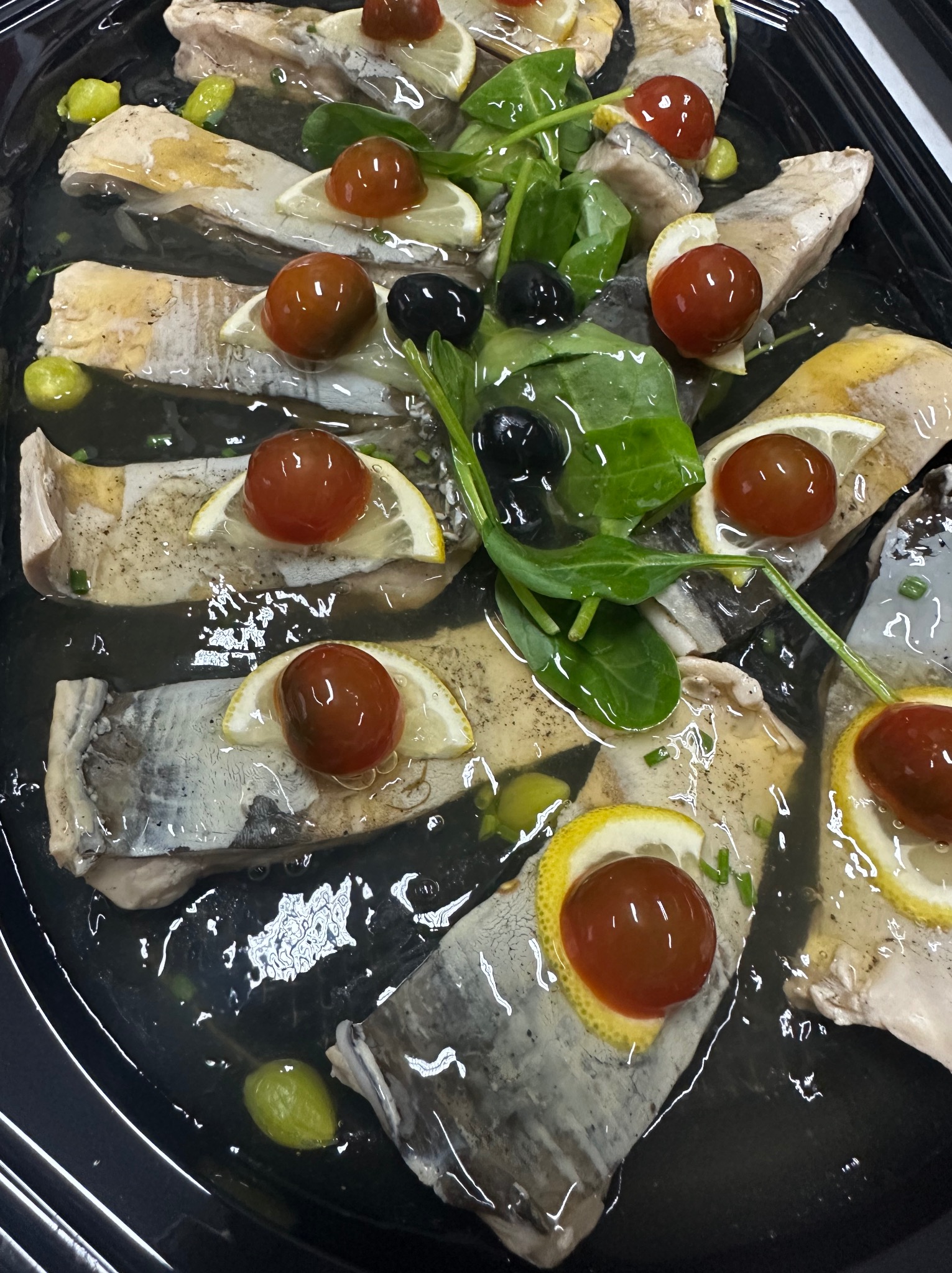 Śledzie w oleju, udekorowane cytryną, pomidorkami cherry, oliwkami i świeżym szpinakiem, ułożone na czarnym talerzu. Gotowa potrawa cateringowa.