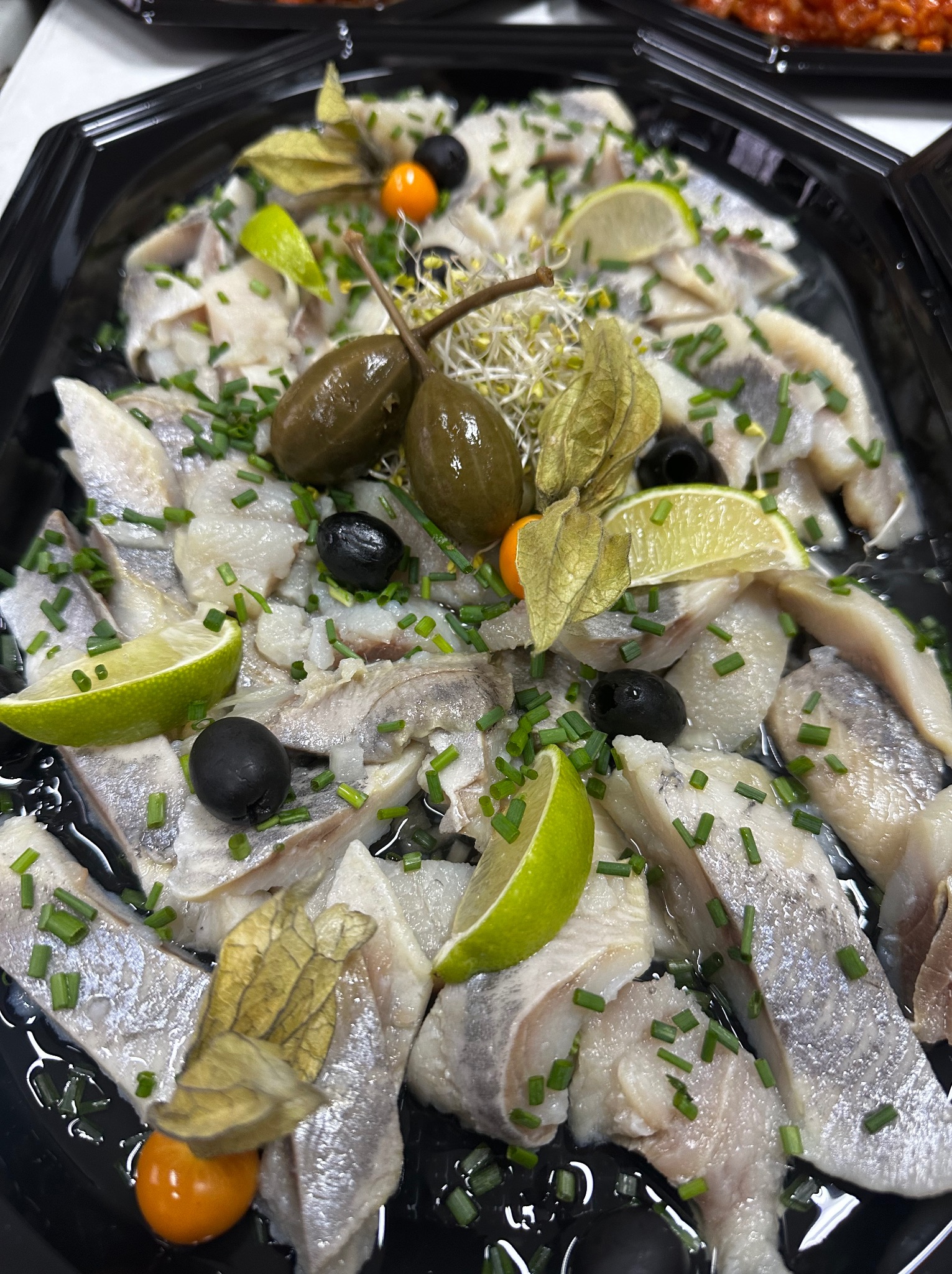 Śledź w oleju, udekorowany kaparami, limonką, czarnymi oliwkami i świeżym szczypiorkiem, podany w czarnym, eleganckim naczyniu cateringowym.