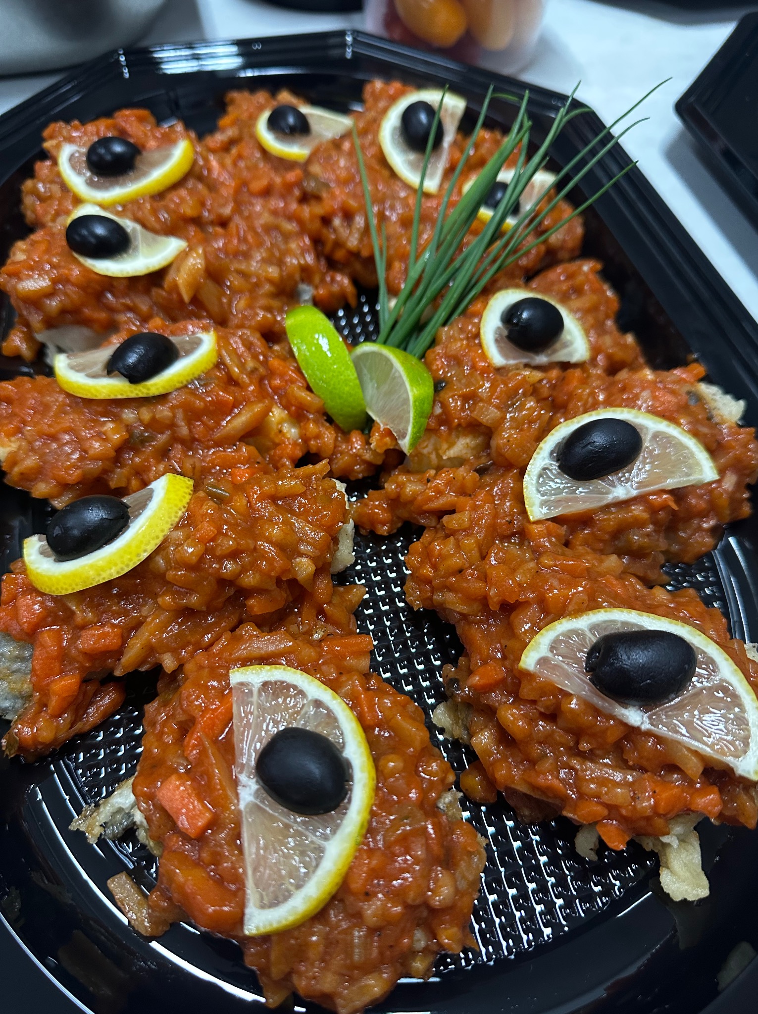 Śledź w pomidorach na czarnym talerzu, udekorowany cytryną, limonką, oliwkami i szczypiorkiem. Elegancka kompozycja potrawy cateringowej.