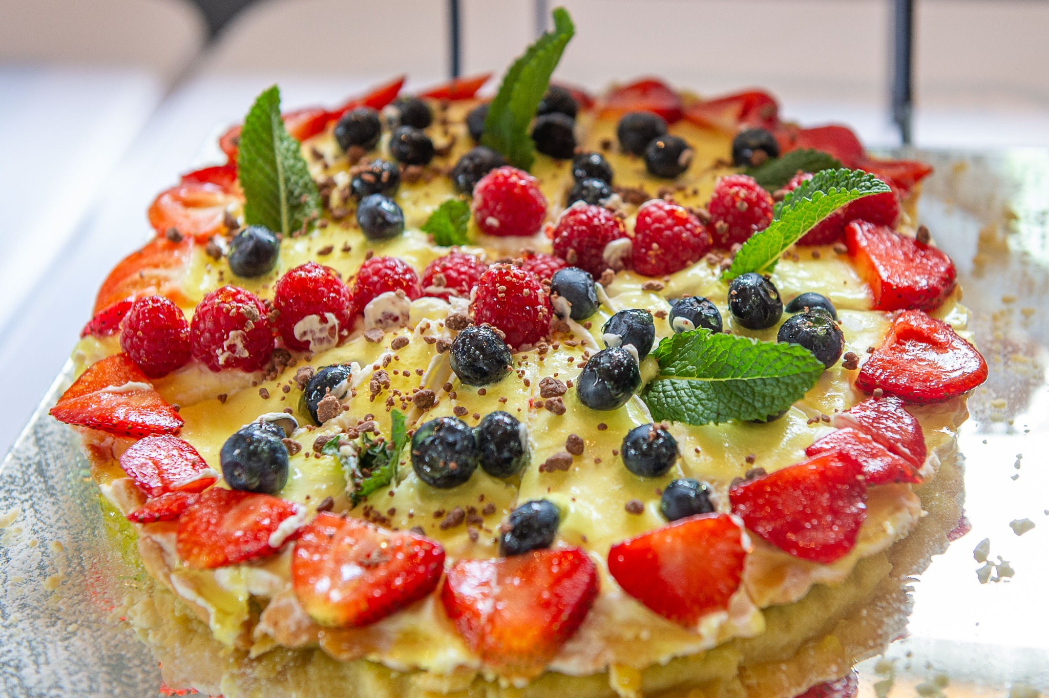 Tarta z kremem, truskawkami, malinami i borówkami, udekorowana listkami mięty i posypana czekoladą, na srebrnej paterze, w jasnym oświetleniu.