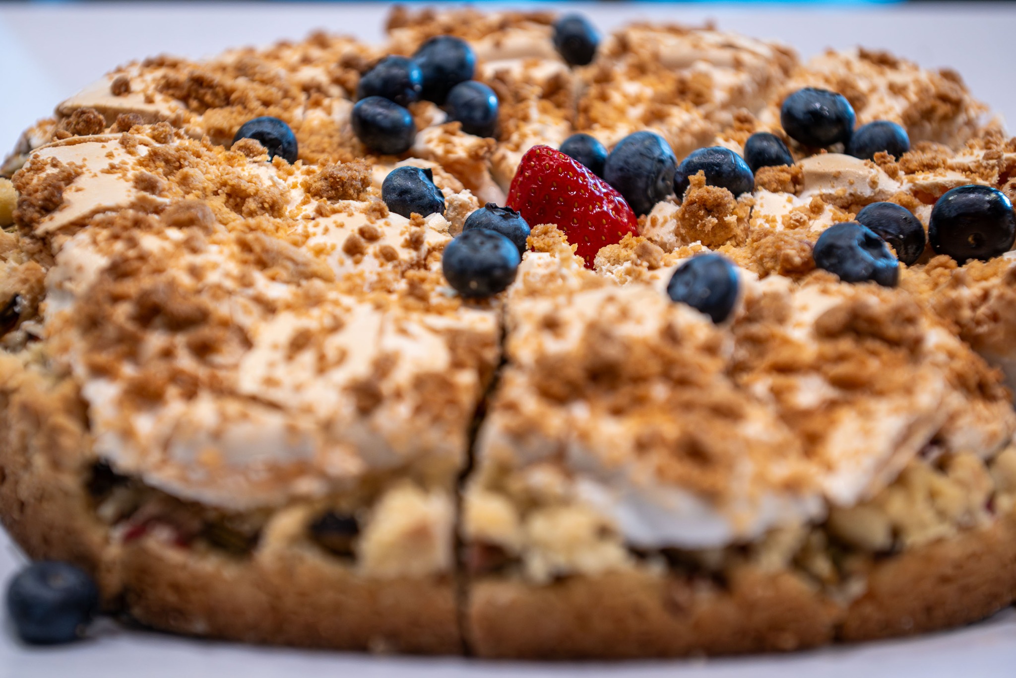 Pleśniak z borówkami i truskawką, widoczny z bliska, z wyraźną strukturą kruszonki i kremu, na białym tle. Delikatne światło podkreśla świeżość owoców.