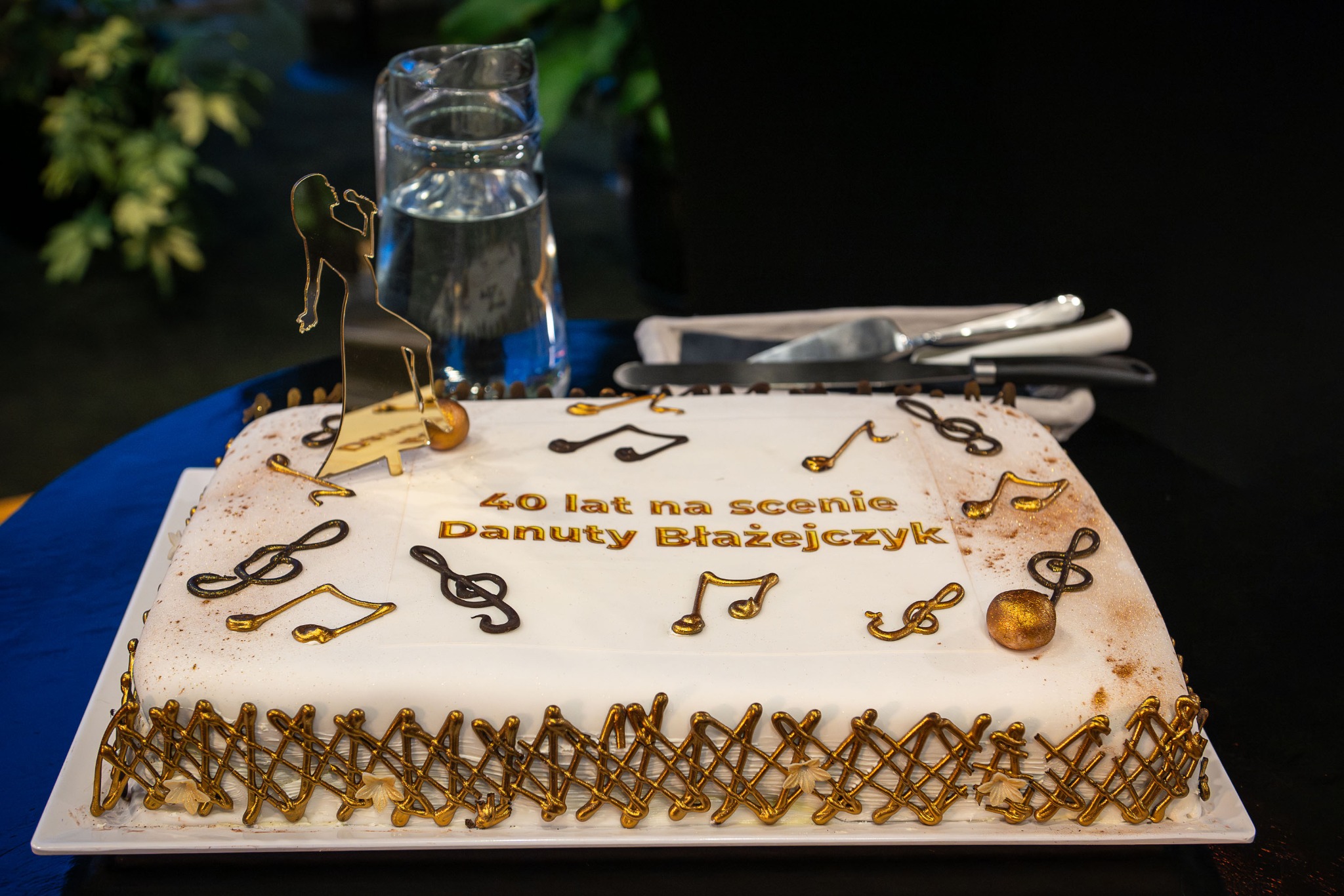 Tort urodzinowy z napisem '40 lat na scenie Danuty Błażejczyk', nutami i figurką piosenkarki, na białym talerzu, obok dzbanek z wodą i sztućce.