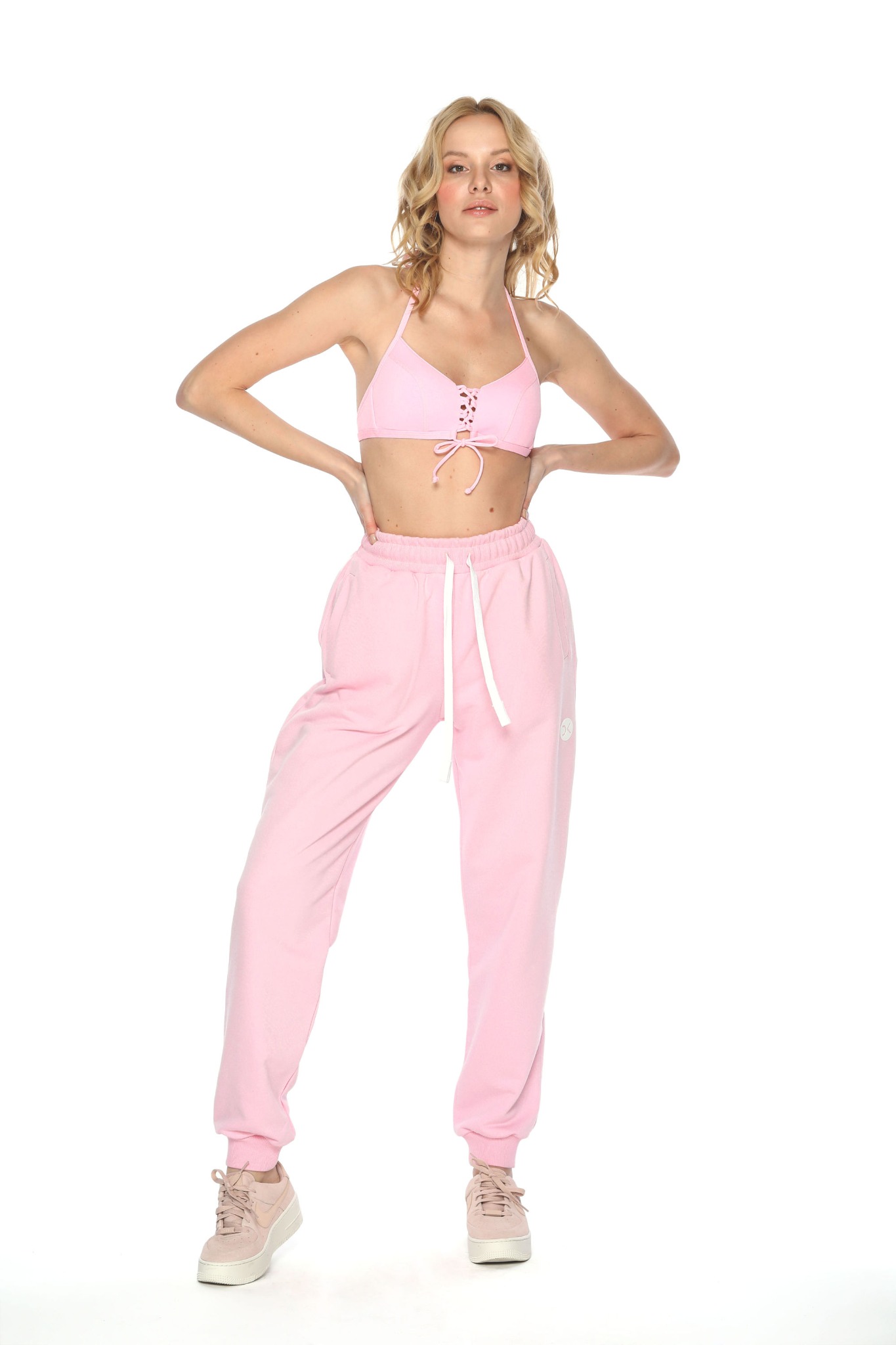 Kobieta pozuje w różowym komplecie: wiązany biustonosz i dresowe spodnie z logo, na białym tle.