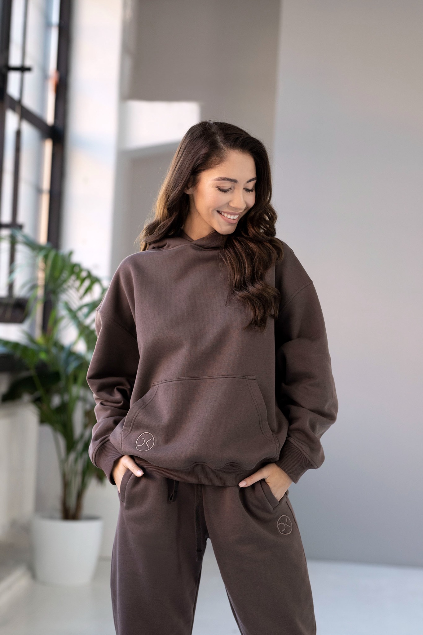 Uśmiechnięta kobieta w brązowym dresie z kapturem i logo na bluzie i spodniach, stojąca na tle okna i rośliny doniczkowej.