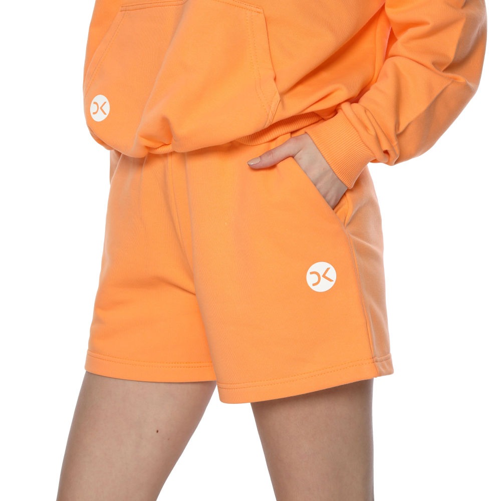 Pomarańczowy komplet dresowy: bluza z kapturem i krótkie spodenki z logo, ręka w kieszeni, widoczne nogi modelki.
