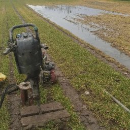 GEORADA Ada Romanowicz - Wiertnica udarowa na błotnistym polu, obok widoczne ślady wierceń i stojąca woda, w tle budynek mieszkalny i drzewa.
